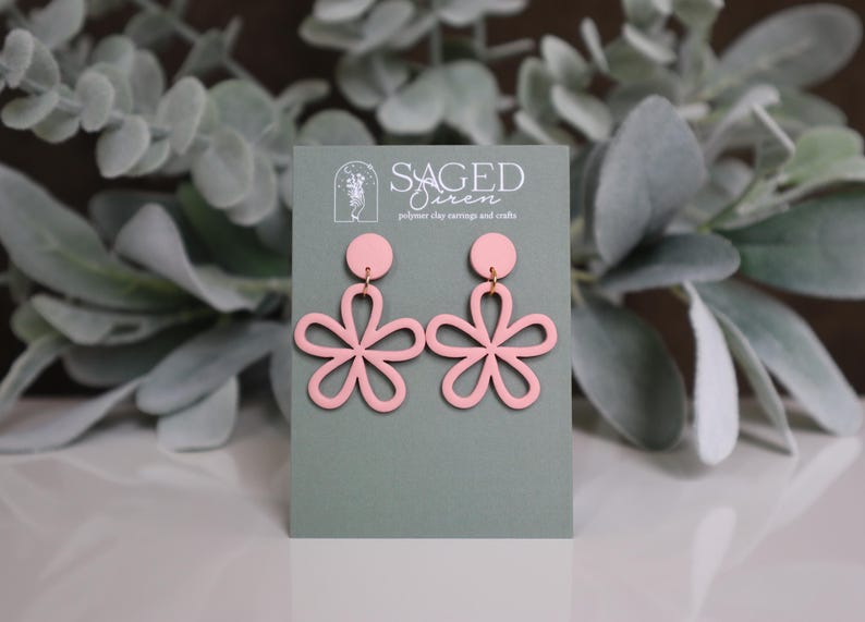 May include: Pink flower-shaped earrings with a circular top and a floral design. The earrings are displayed on a gray card with the text "SAGED Siren polymer clay earrings and crafts". The background features green foliage.