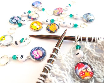 May include: A set of colorful owl-themed stitch markers with numbers and beads. The markers are attached to knitting needles and a white knitted fabric.