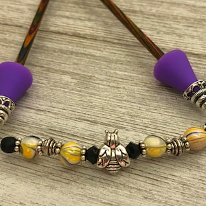 May include: A pair of purple hair sticks with black and yellow beads and a silver bee charm. The sticks are made of wood with a multi-colored swirl pattern.