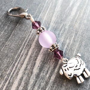 Puede incluir: Un amuleto de plata con un diseño de oveja, unido a un llavero de plata con una cuenta morada y detalles de cristal.