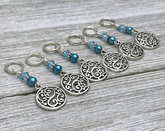 May include: Six blue and silver stitch markers with a decorative swirl design. Each marker has a blue bead and a crystal.