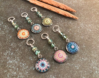 May include: Six colorful stitch markers with mandala designs. Each marker has a silver ring and a green glass bead. The markers are arranged on a brown textured surface.