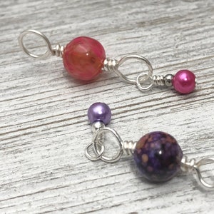 Puede incluir: Dos juegos de dijes de joyería con una mezcla de cuentas moradas, rosadas y transparentes con detalles de alambre plateado.