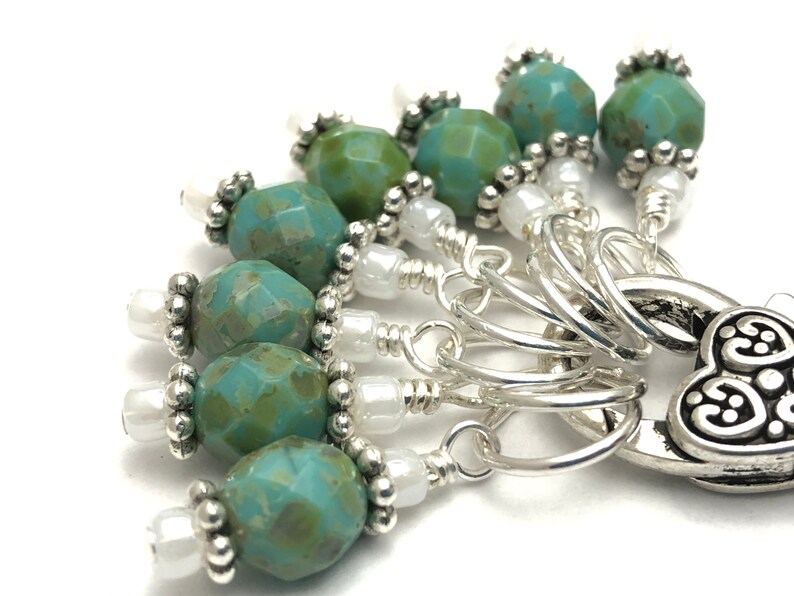 May include: A set of 7 stitch markers with a heart-shaped charm. The markers are silver with turquoise and white beads.