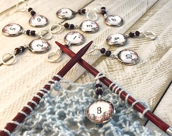 May include: Ten numbered stitch markers with floral designs and purple beads on silver rings. The markers are on a wooden surface with two knitting needles and a blue knitted fabric.