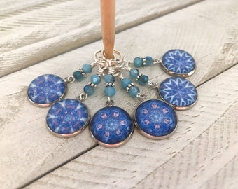 May include: Six blue and white mandala stitch markers with silver rings and blue crystal beads. The stitch markers are hanging from a wooden knitting needle.