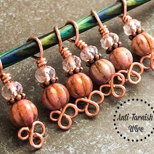 May include: Six copper wire stitch markers with pink and white striped beads and clear crystal beads. The markers are attached to a wooden knitting needle. "Anti-Tarnish Wire" is written on the bottom of the image.
