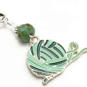 May include: A green and gold charm of a yarn ball with a tail of yarn. The charm is attached to a silver chain with a green and blue glass bead.