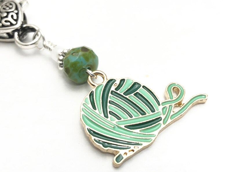 May include: A green and gold charm of a yarn ball with a tail of yarn. The charm is attached to a silver chain with a green and blue glass bead.