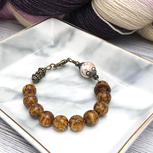 Pode incluir: Um bracelete de contas marrons e brancas com um fecho de latão e uma conta de cerâmica branca com um desenho floral.