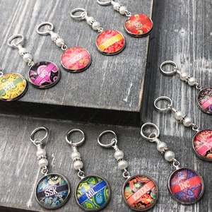 Puede incluir: Diez marcadores de punto coloridos con cuentas blancas y anillos plateados. Cada marcador presenta un diseño de patrón diferente con una abreviatura de tejido en texto blanco.