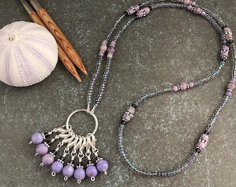 May include: A purple and silver beaded necklace with a silver ring and multiple purple beads with silver clips hanging from it. The necklace is on a gray surface with a purple sea urchin and two wooden knitting needles.