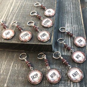 Puede incluir: Diez marcadores de punto de plata con cuentas marrones y discos redondos con estampado floral. Cada disco tiene una abreviatura de tejido diferente escrita en texto negro. Las abreviaturas son: SKP, P2TOG, PURL, K2TOG, YO, SSK, M1L, SK2P, M1R y KNIT.