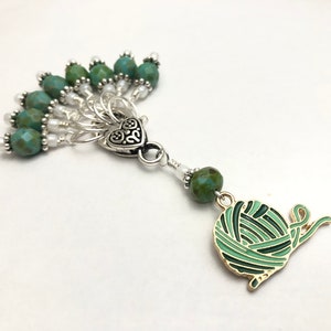 May include: A green and gold yarn ball stitch marker with a heart charm and a fan of green and white beads.