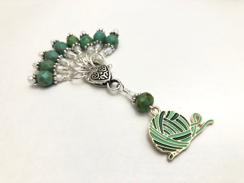 May include: A green and gold yarn ball stitch marker with a heart charm and a fan of green and white beads.