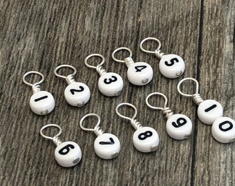 May include: Ten white stitch markers with black numbers 1 through 10. The markers are made of plastic and have a silver wire loop.