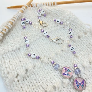 Puede incluir: Marcadores de punto morado y plateado con números del 0 al 9 y dijes de mariposa. Los marcadores están enhebrados en una cadena y se utilizan para contar los puntos en el tejido de punto.