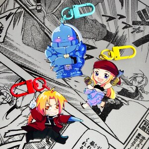 May include: Three acrylic keychains featuring anime-style characters. One is a blue armored figure with a light blue clasp, one is a blonde character in a red coat with a red clasp, and one is a blonde character with a wrench and a yellow clasp.
