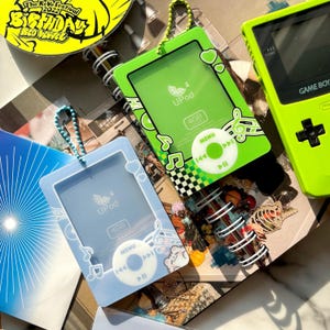 Puede incluir: Primer plano de dos fundas UPod, una verde y otra azul claro, con temática musical. La funda verde tiene un patrón de tablero de ajedrez y notas musicales. La funda azul claro tiene un diseño de corazón. Ambas fundas tienen el texto "UPod" y "4GB". También se ve una Game Boy verde.