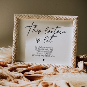 Op de afbeelding: Een wit houten frame met een witte achtergrond en zwarte tekst die "This lantern is lit. In loving memory of those who are forever present in our hearts. We love and miss you!" luidt. Het frame ligt op een bruine stoffen ondergrond.