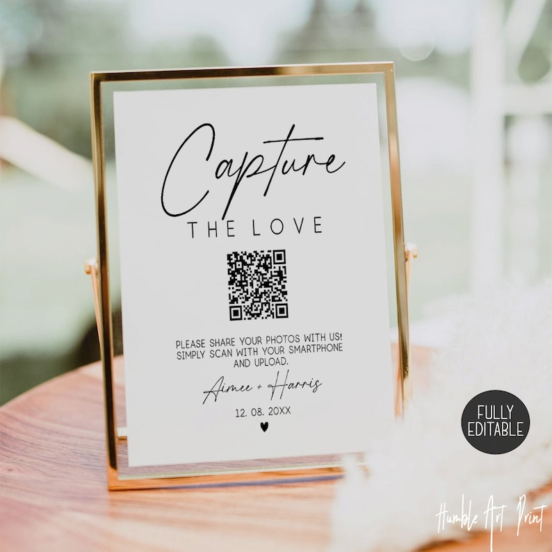 May include: A white sign with a gold frame, featuring the text "Capture the Love" in black cursive font. A black and white QR code is displayed below the text, with instructions to scan the code with a smartphone to share photos. The sign also includes the names "Aimee + Harris" and the date "12. 08. 20XX" in black cursive font. The text "FULLY EDITABLE" is displayed in black on a white background in the bottom right corner of the sign.