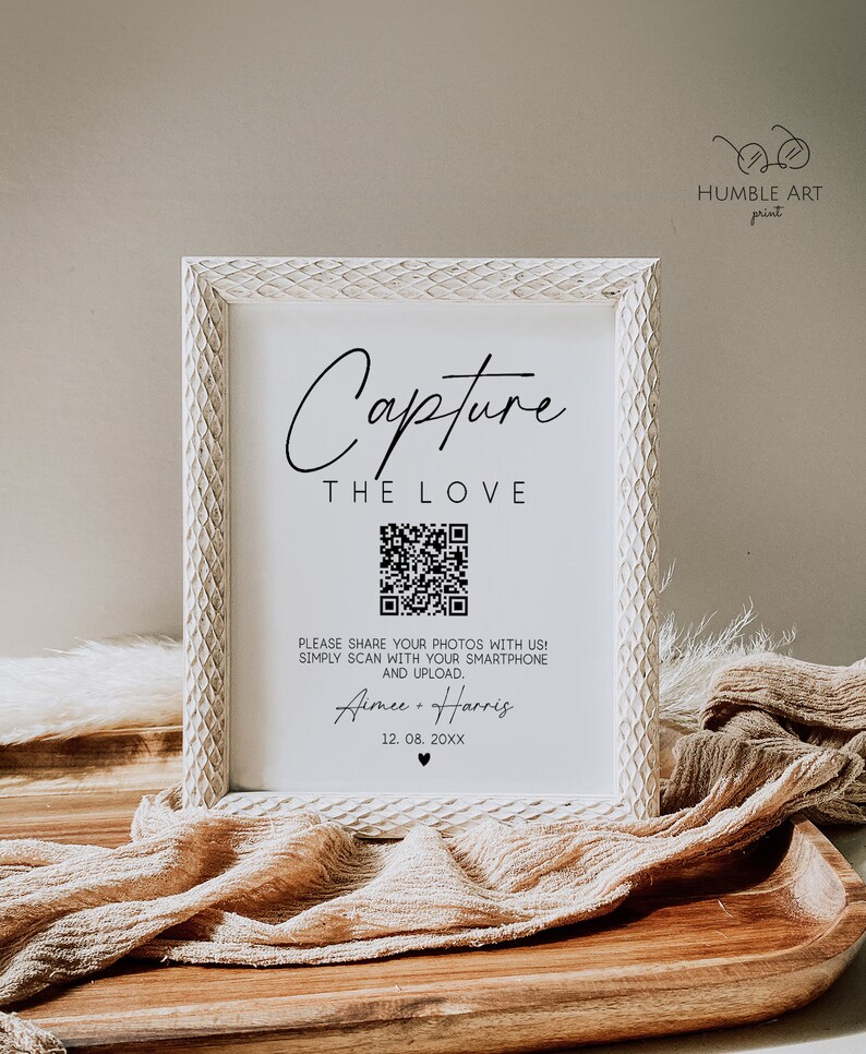 May include: White framed sign with the text "Capture the Love" and a QR code. The text below the QR code reads "Please share your photos with us! Simply scan with your smartphone and upload. Aimee & Harris 12.08.20XX".