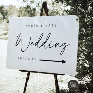 May include: White wedding sign with black text that reads "Amber & Nate Wedding This Way" with a black arrow pointing to the right. The sign is on a wooden easel.