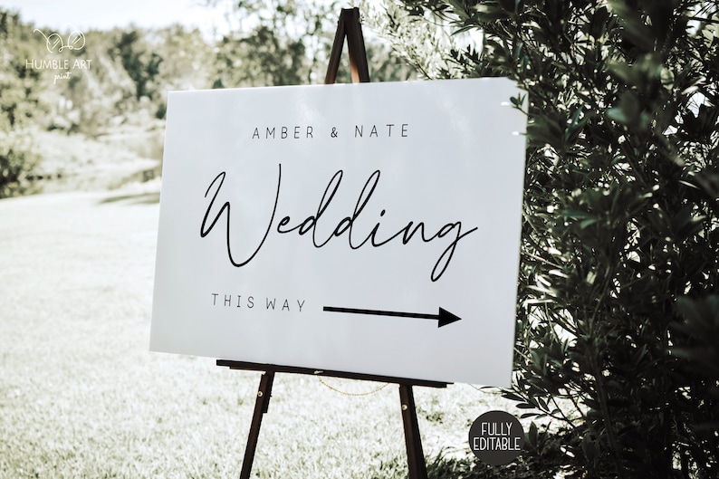 May include: White wedding sign with black text that reads "Amber & Nate Wedding This Way" with a black arrow pointing to the right. The sign is on a wooden easel.