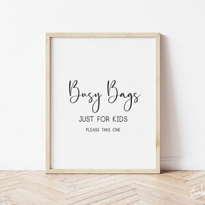 May include: A framed print with the words "Busy Bags JUST FOR KIDS PLEASE TAKE ONE" in black script on a white background. The frame is a light wood color, and the background is a white wall with a wooden floor.