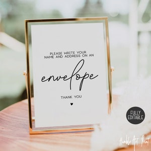 May include: A white sign with black text in a gold frame. The sign says "Please write your name and address on an envelope. Thank you." with a small heart symbol below the text. The sign is sitting on a wooden surface.