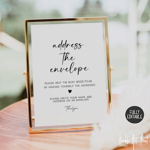 May include: A white sign with black text that reads "address the envelope". The sign also includes instructions for guests to write their name and address on an envelope for the bride-to-be. The sign is framed in a gold frame and is sitting on a wooden table.