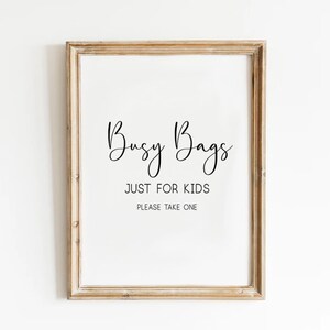 May include: A framed sign with the words "Busy Bags" in a cursive font, followed by "JUST FOR KIDS" and "PLEASE TAKE ONE" in a simple font. The frame is a light brown color.