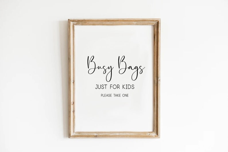 May include: A framed sign with the words "Busy Bags" in a cursive font, followed by "JUST FOR KIDS" and "PLEASE TAKE ONE" in a simple font. The frame is a light brown color.