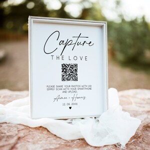 May include: White sign with black text that reads "Capture the Love". A black and white QR code is below the text. The text below the QR code reads "Please share your photos with us! Simply scan with your smartphone and upload. Aimee + Harris 12.08.20XX".