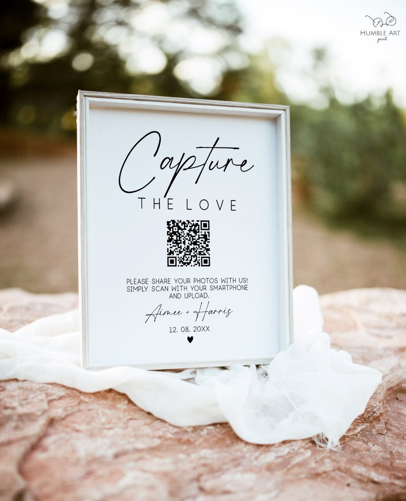 May include: White sign with black text that reads "Capture the Love". A black and white QR code is below the text. The text below the QR code reads "Please share your photos with us! Simply scan with your smartphone and upload. Aimee + Harris 12.08.20XX".
