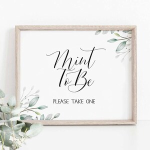 May include: A white sign with a wooden frame and green foliage in the corners. The sign says "Mint To Be" in black cursive lettering and "Please Take One" in smaller black lettering.
