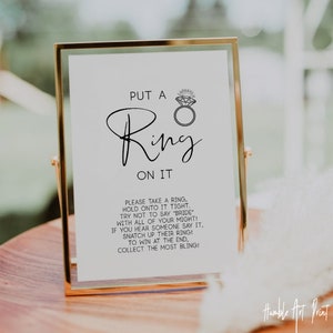 May include: A white sign with black text in a gold frame that reads "Put a Ring on It". The sign also includes instructions for a game: "Please take a ring, hold onto it tight. Try not to say "Bride" with all of your might! If you hear someone say it, snatch up their ring! To win at the end, collect the most bling!"