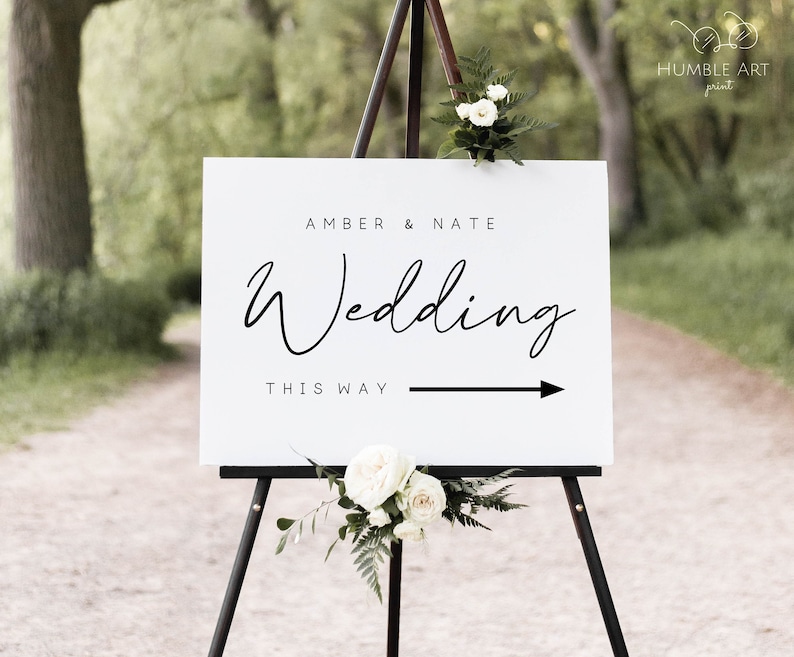 May include: White wedding sign with black text that reads "Amber & Nate Wedding This Way" with a black arrow pointing to the right. The sign is on a black easel with white flowers.