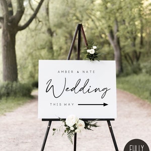 May include: White wedding sign with black text that reads "Amber & Nate Wedding This Way" with a black arrow pointing to the right. The sign is on a black easel in a forest setting.