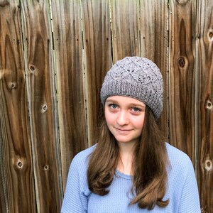 May include: A gray knitted beanie with a diamond pattern. The beanie is worn by a person with long brown hair and a blue shirt.