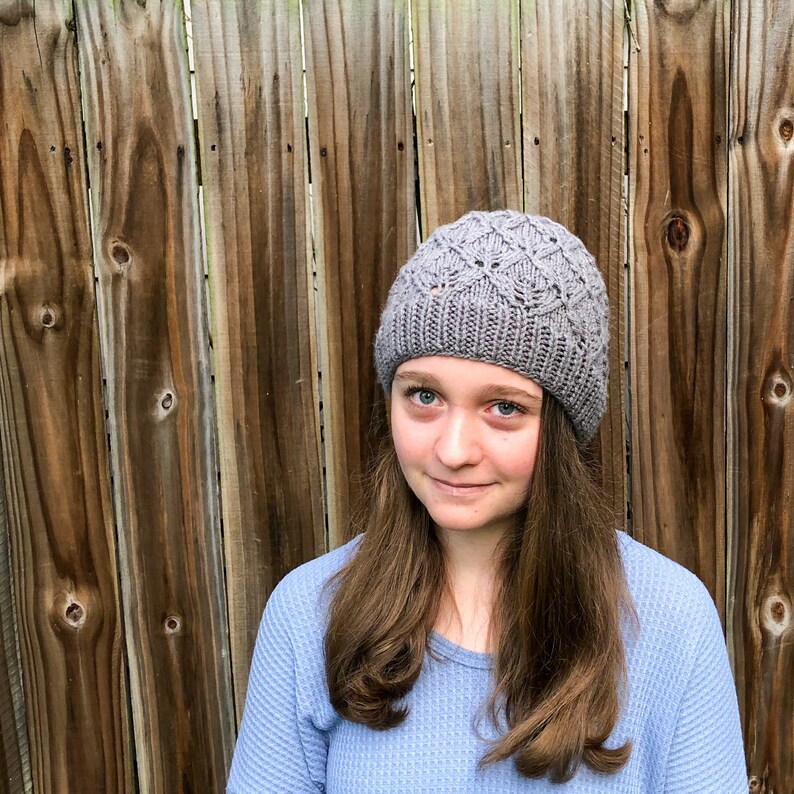 May include: A gray knitted beanie with a diamond pattern. The beanie is worn by a person with long brown hair and a blue shirt.