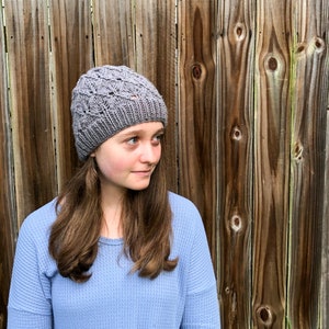 May include: A woman wearing a gray knitted hat with a lacy pattern and a light blue long-sleeved shirt. She is standing in front of a wooden fence.