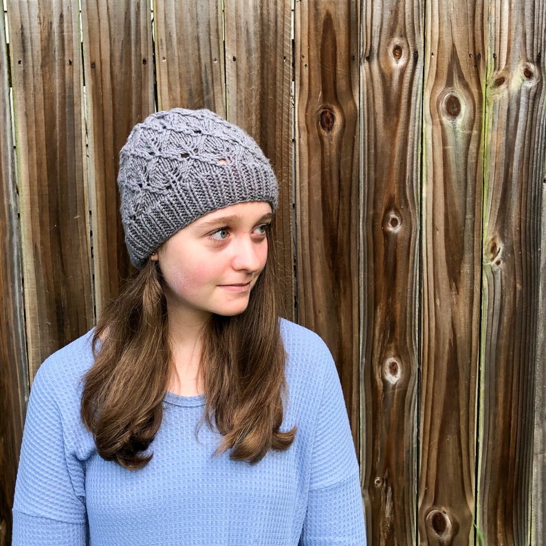May include: A woman wearing a gray knitted hat with a lacy pattern and a light blue long-sleeved shirt. She is standing in front of a wooden fence.