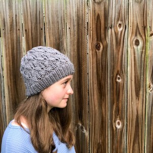 May include: A gray knitted hat with a lacy pattern. The hat is worn by a person with long brown hair.
