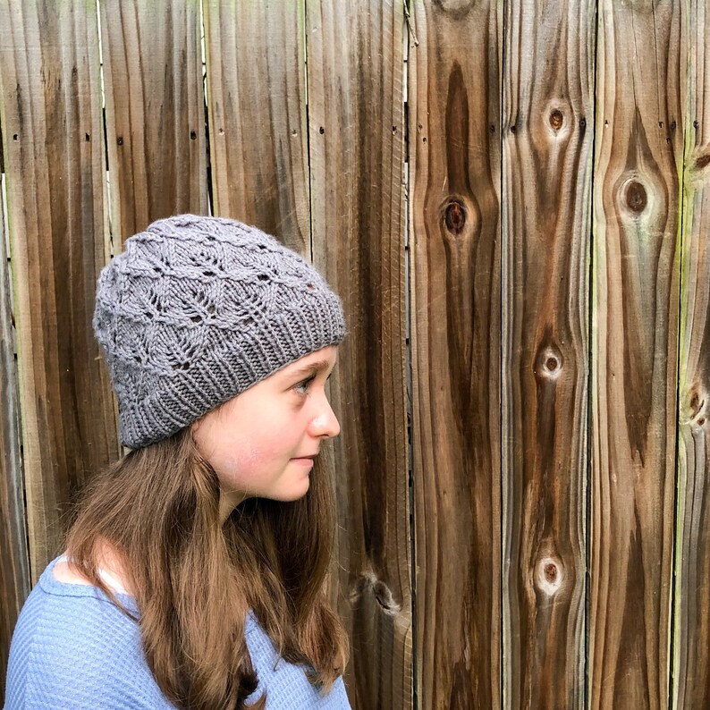 May include: A gray knitted hat with a lacy pattern. The hat is worn by a person with long brown hair.