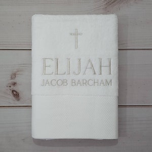 May include: White personalised towel with embroidered cross, the name "ELIJAH", and the name "JACOB BARCHAM" in silver thread. The bottom section of the towel has a textured pattern.