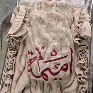 May include: A newborn baby wrapped in a beige knitted blanket with ruffled edges. The blanket has a maroon Arabic script design. The baby is lying in a clear bassinet.
