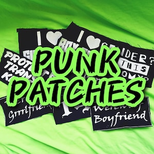 May include: A collection of rectangular black fabric patches with white printed text and graphics. Visible phrases include I LOVE BOY GENDER, PROTECT TRANS KIDS, GRRLFRIEND, and WEREWOLF BOYFRIEND. The patches are arranged in a cluster on a bright lime green surface.