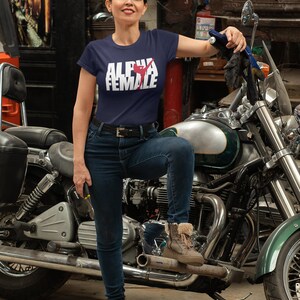May include: A navy blue t-shirt with the text "ALPHA FEMALE" in white block letters and a red graphic. The woman in the image is wearing blue jeans and brown boots, leaning on a motorcycle.