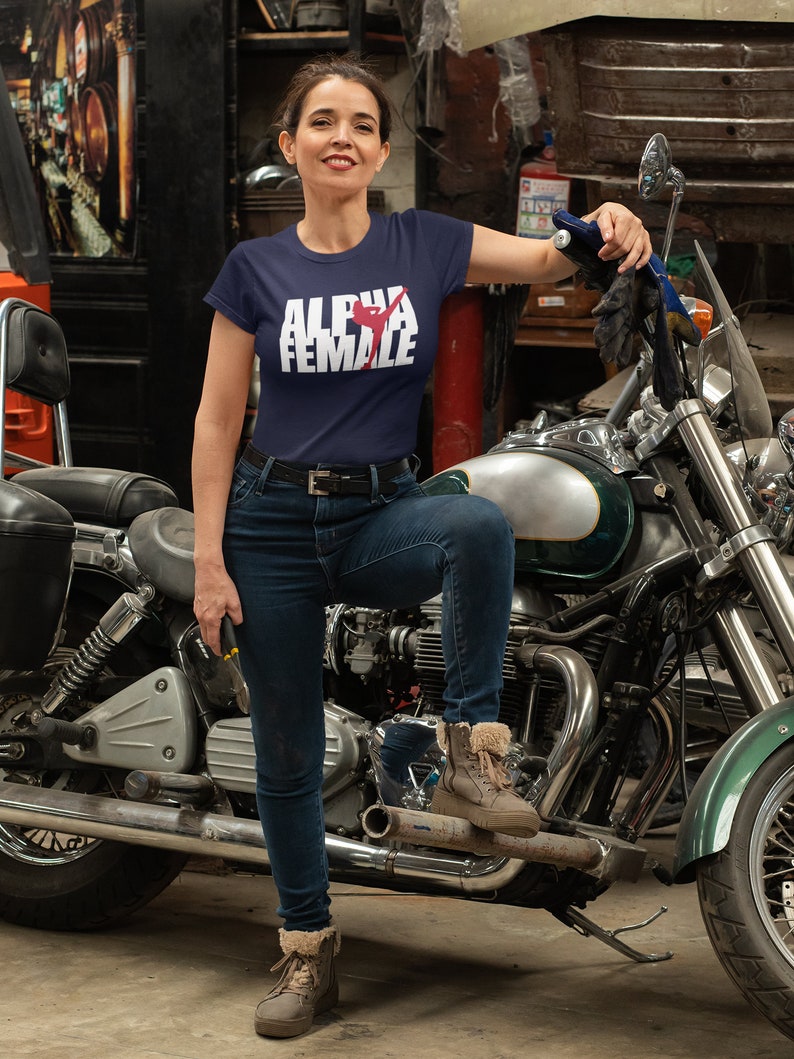 May include: A navy blue t-shirt with the text "ALPHA FEMALE" in white block letters and a red graphic. The woman in the image is wearing blue jeans and brown boots, leaning on a motorcycle.